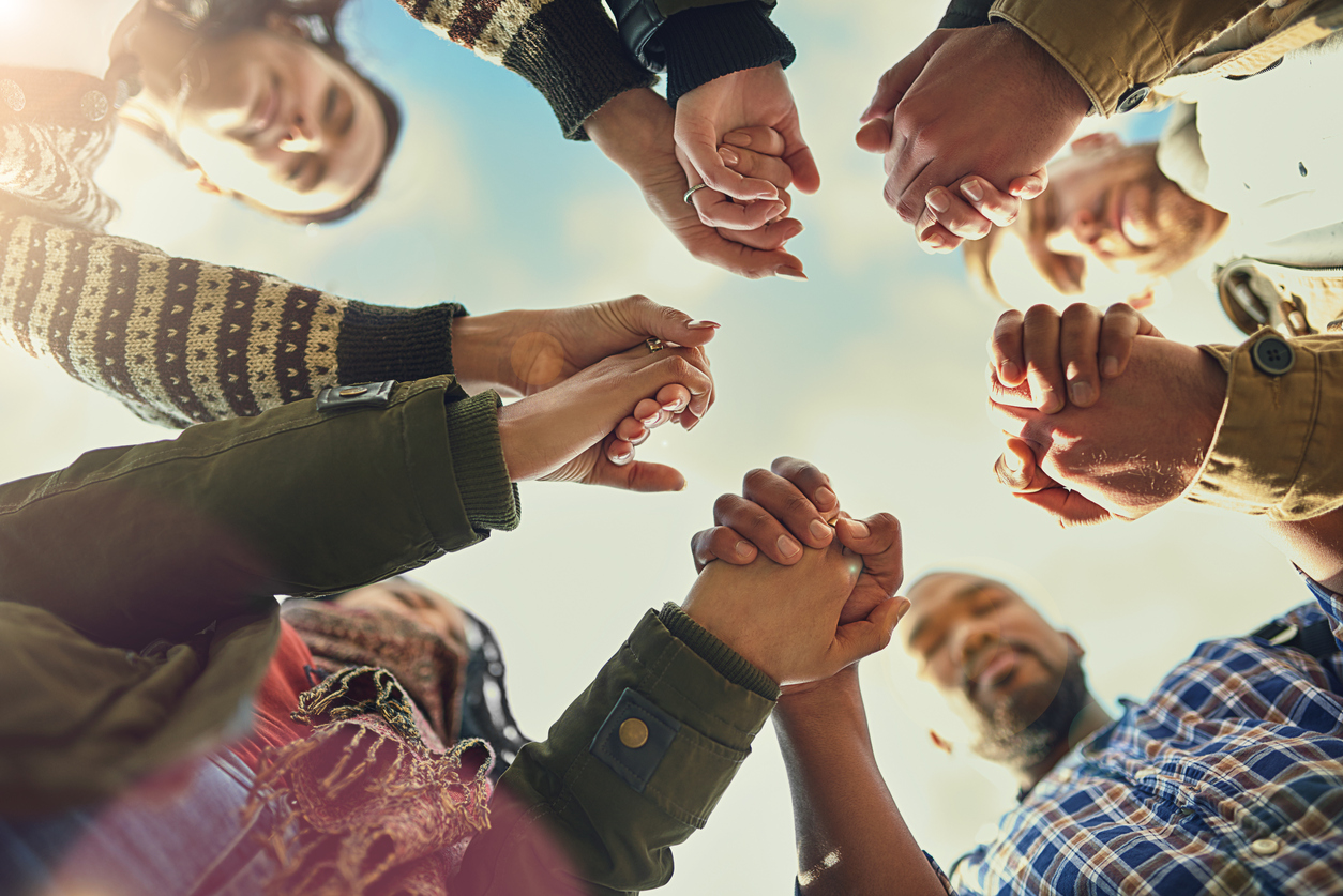 A low-angle of a group of diverse people in a circle holding hands, in prayer, symbolizing unity and community.